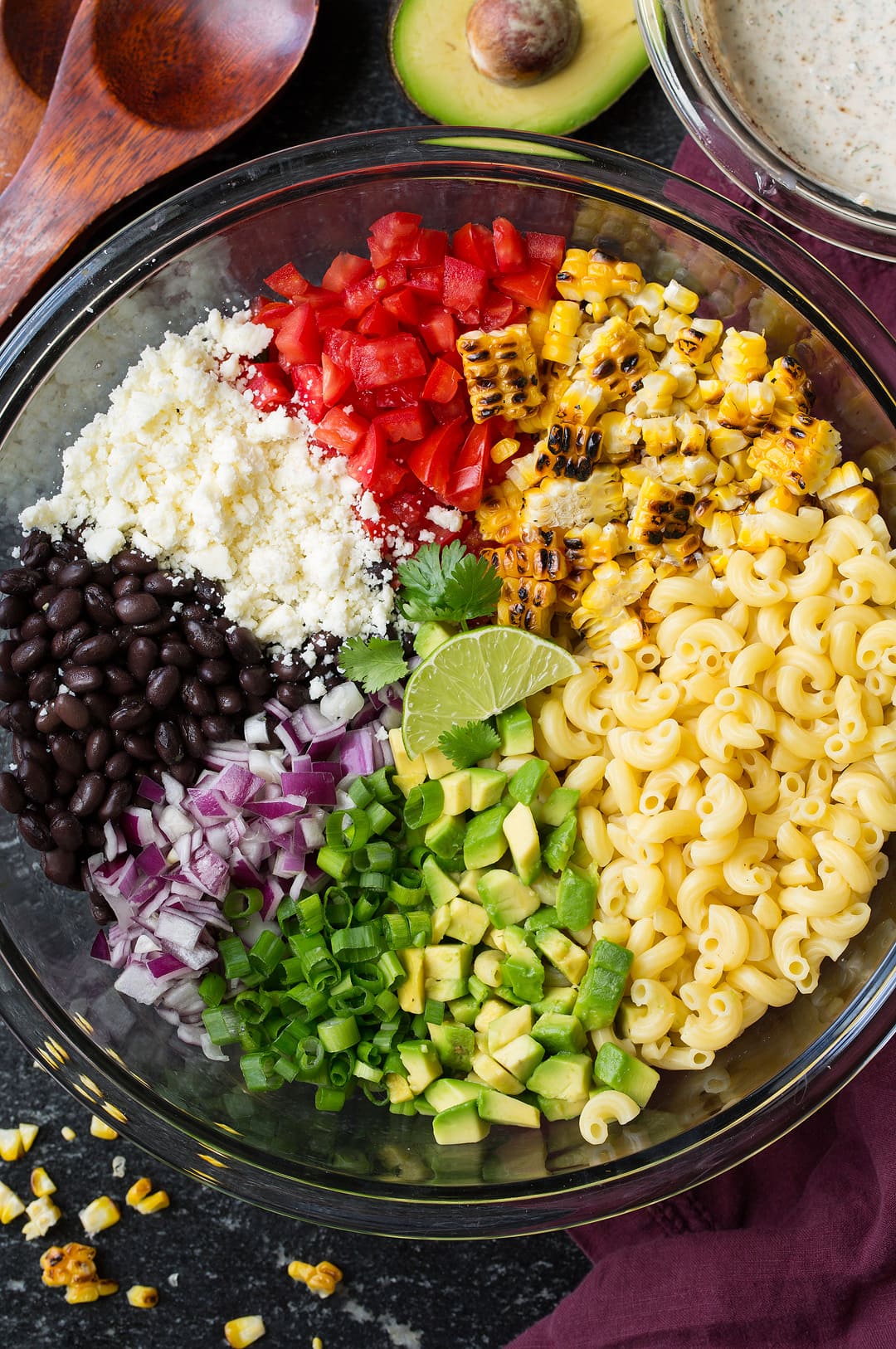 Mexican macaroni salad ingredients in a large glass bowl before tossing. Ingredients include macaroni, charred corn, black beans, avocado, tomato, green onion, red onion, and cotija. 