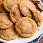 Image of snickerdoodles cookies shown from the side layered together on a white plate with cinnamon sticks in the background. Plate is resting on a striped blue cloth and wooden platter.