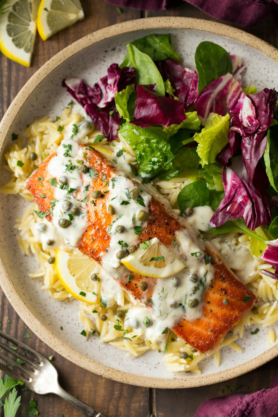 Salmon cooking in skillet with creamy lemon sauce and capers. Shown here on a single serving plate with orzo and a side salad.