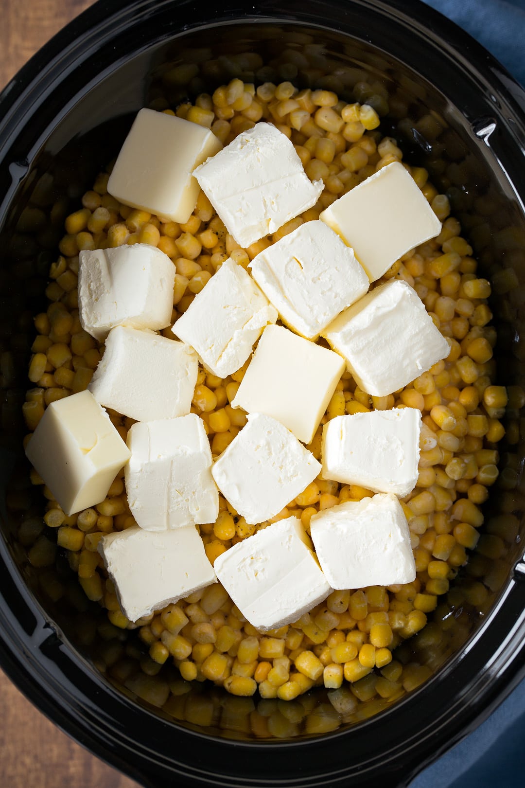 Creamed Corn in a slow cooker. A layer of corn is dotted with cubes of cream cheese before cooking.