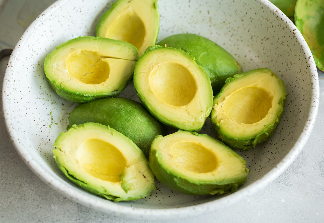 Showing how to make guacamole, adding eight avocado halves to a large white ceramic mixing bowl.