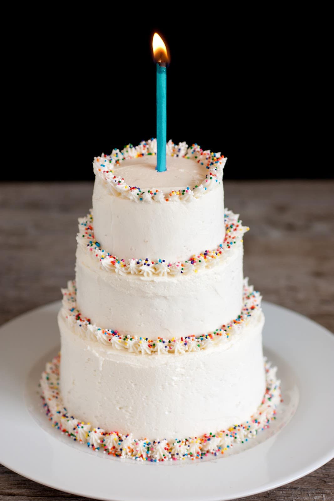 Three tiered cake covered with the best vanilla buttercream frosting. Cake edges are garnished with multi colored sprinkles and there's one lit candle atop the cake.