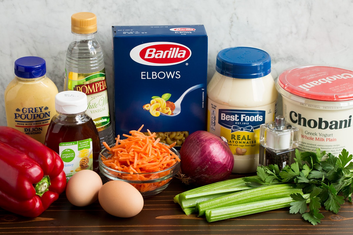 Ingredients that are in macaroni salad shown here. Includes mayonnaise, Greek yogurt, dijon mustard, honey, eggs, carrots, bell pepper, celery, parsley, vinegar, salt and pepper.