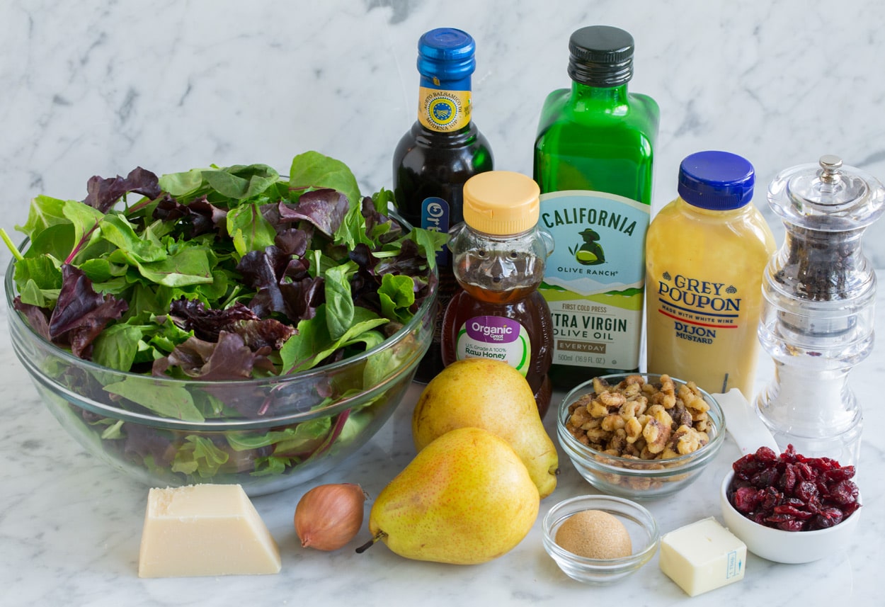 Ingredients for pear salad sitting on marble surface. Includes spring greens, pears, parmesan, olive oil, balsamic vinegar, butter, pecans, cranberries, mustard, honey, brown sugar.