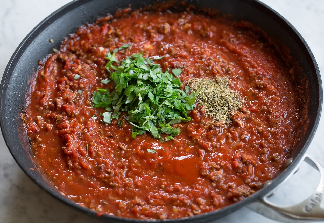 Adding basil and oregano to beefy tomato spaghetti sauce in pan.