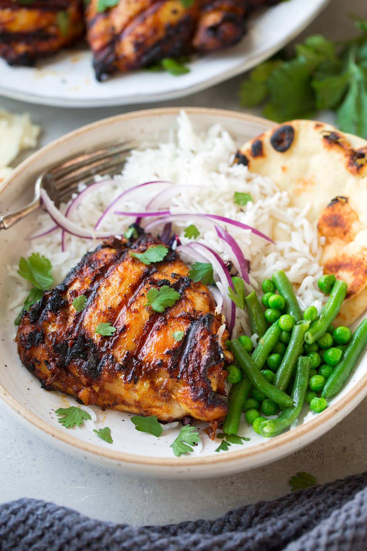 Grilled Tandoori Chicken in a shallow serving bowl with a side of rice, naan, beans and peas. Garnished with red onion and cilantro.