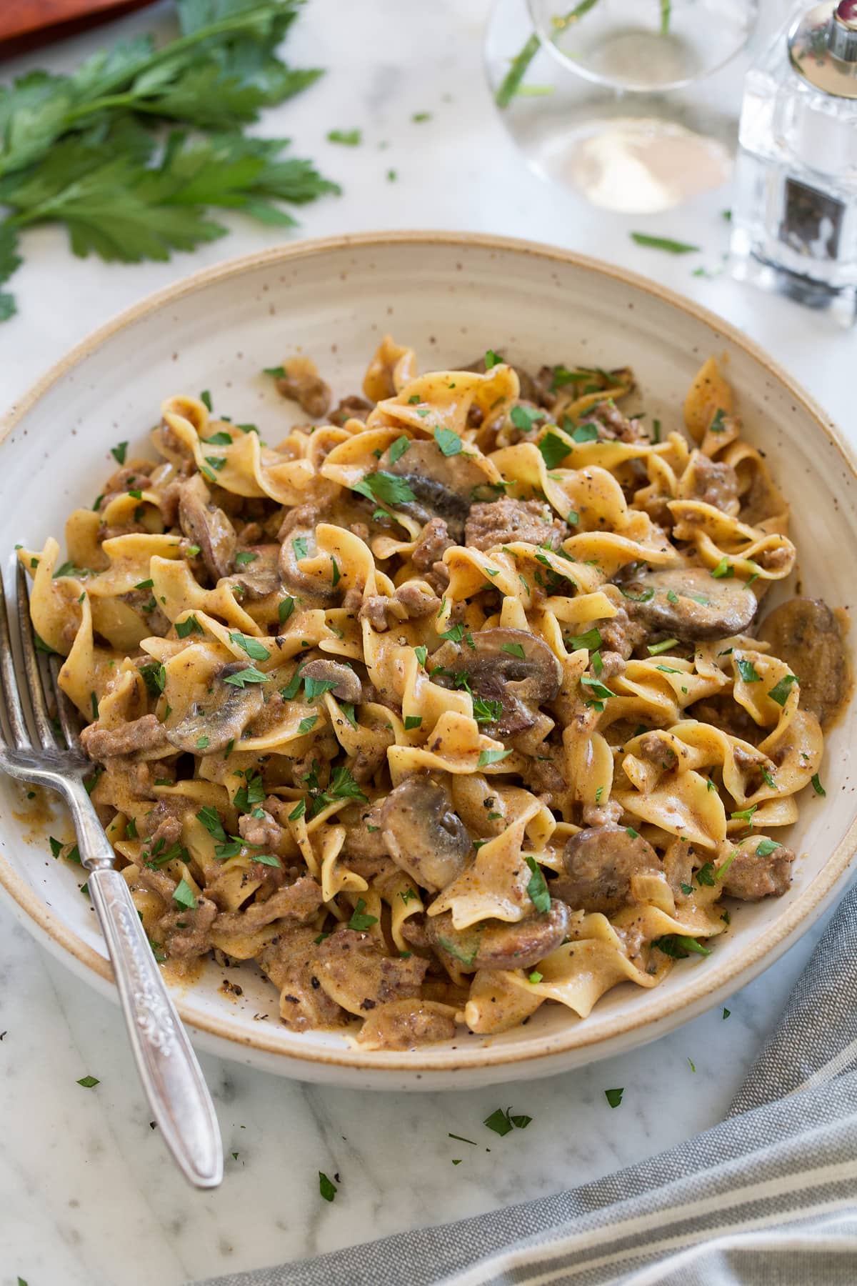 One Pot Easy Beef Stroganoff shown in a cream pasta bowl with a fork to the side. Bowl is sitting on a marble surface, parsley and a salt shaker are in the background.