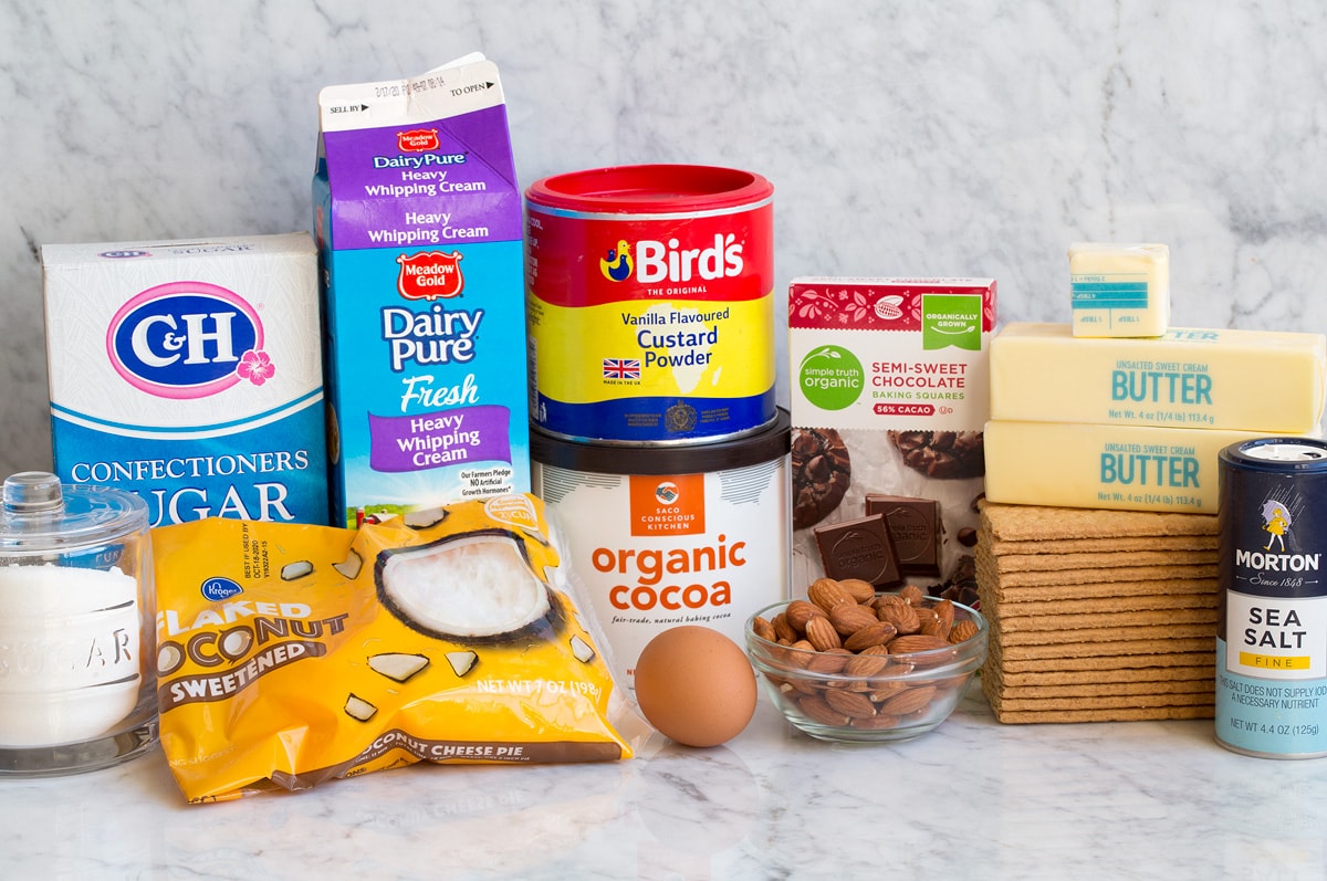 Image of ingredients used to make Nanaimo Bars. Includes sugar, coconut, powdered sugar, cream, custard powder, cocoa powder, egg, almonds, chocolate, graham crackers, butter, and salt.