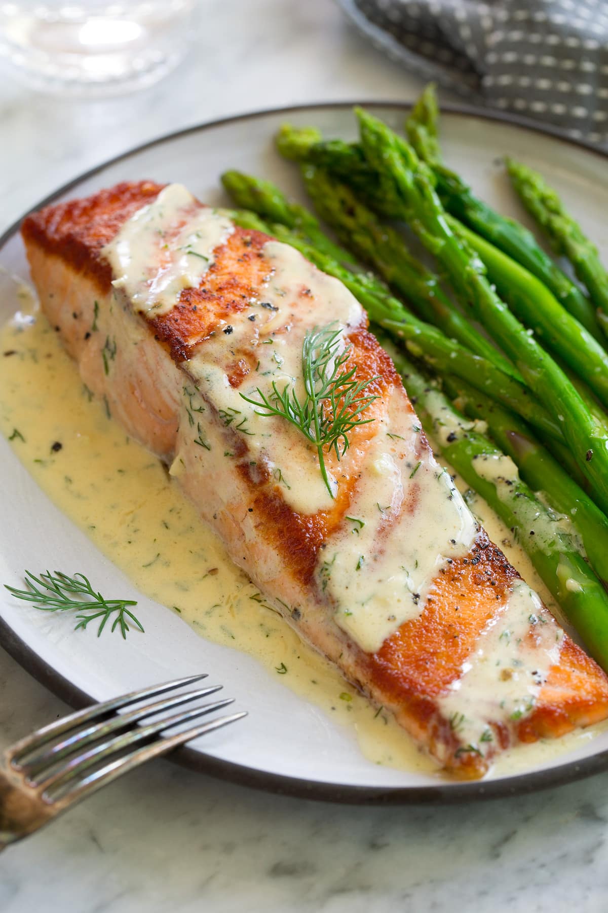 A single pan seared salmon fillet on a white plate. Salmon is drizzled with a creamy mustard sauce and sprinkled with dill. It's served with a side of asparagus.