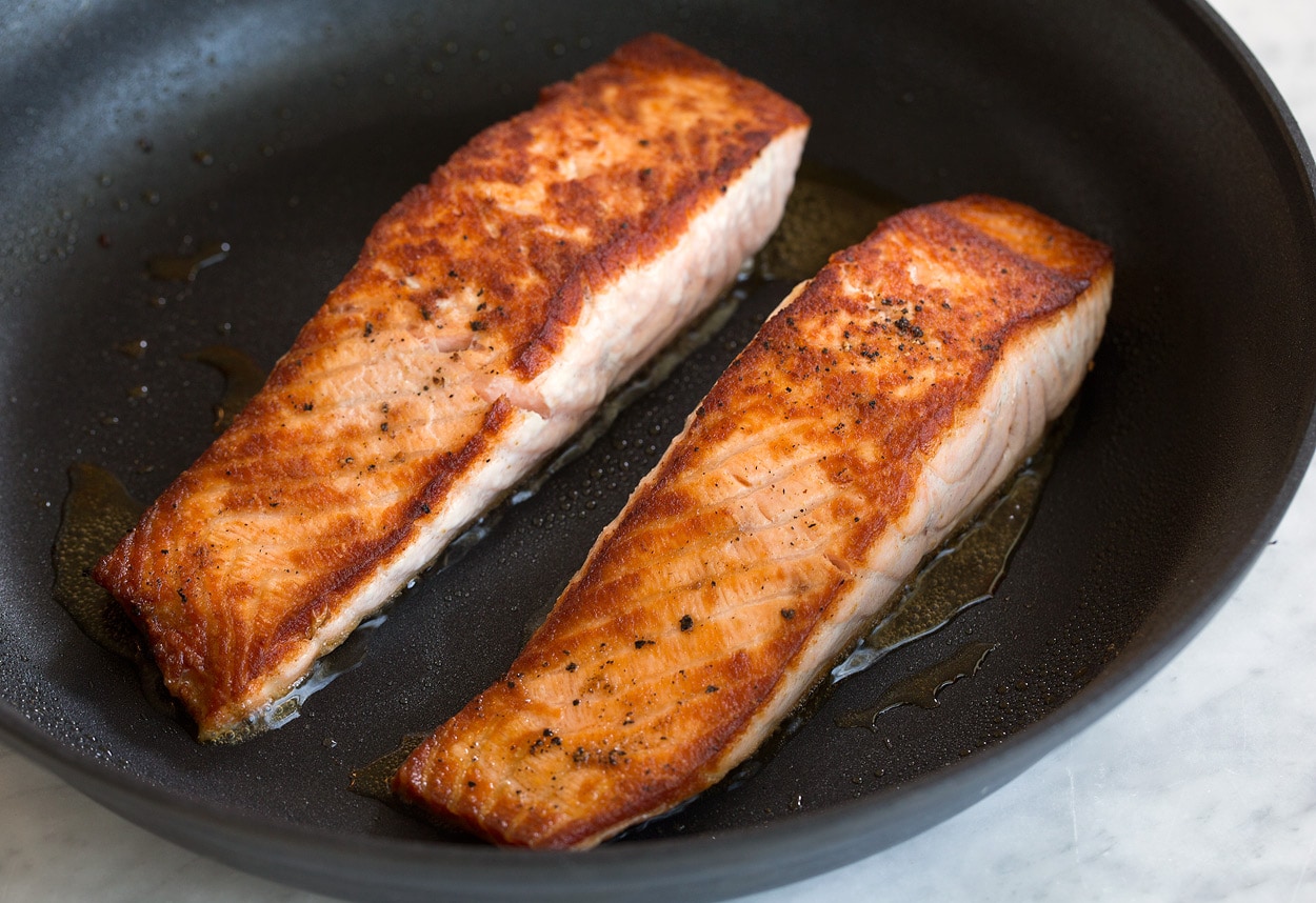 Two golden brown pan seared salmon fillets shown here in large non-stick skillet.