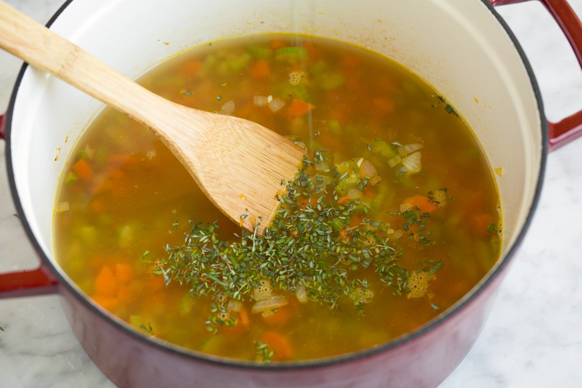 Adding broth and fresh herbs to soup mixture in pot.