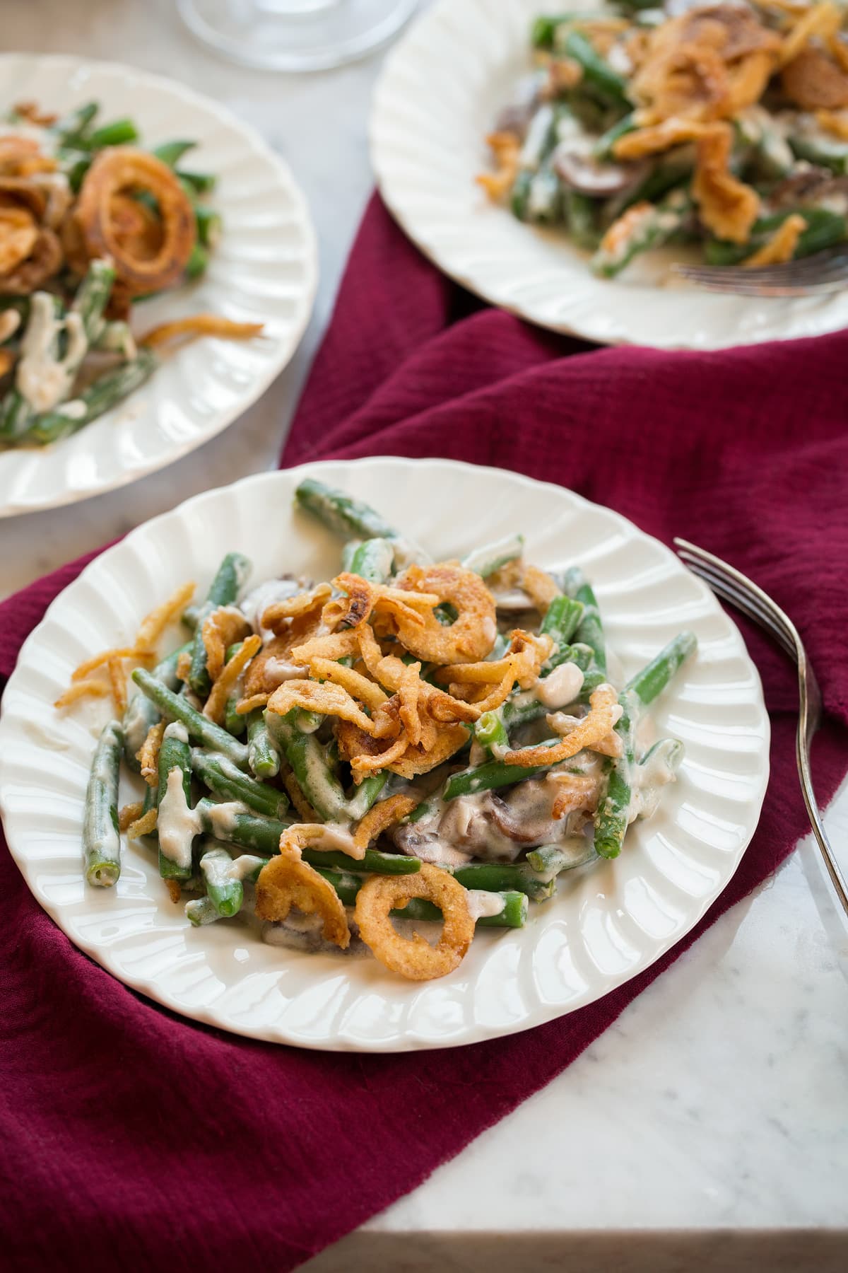 Three individual servings of Green Bean Casserole on white scalloped salad plates. 