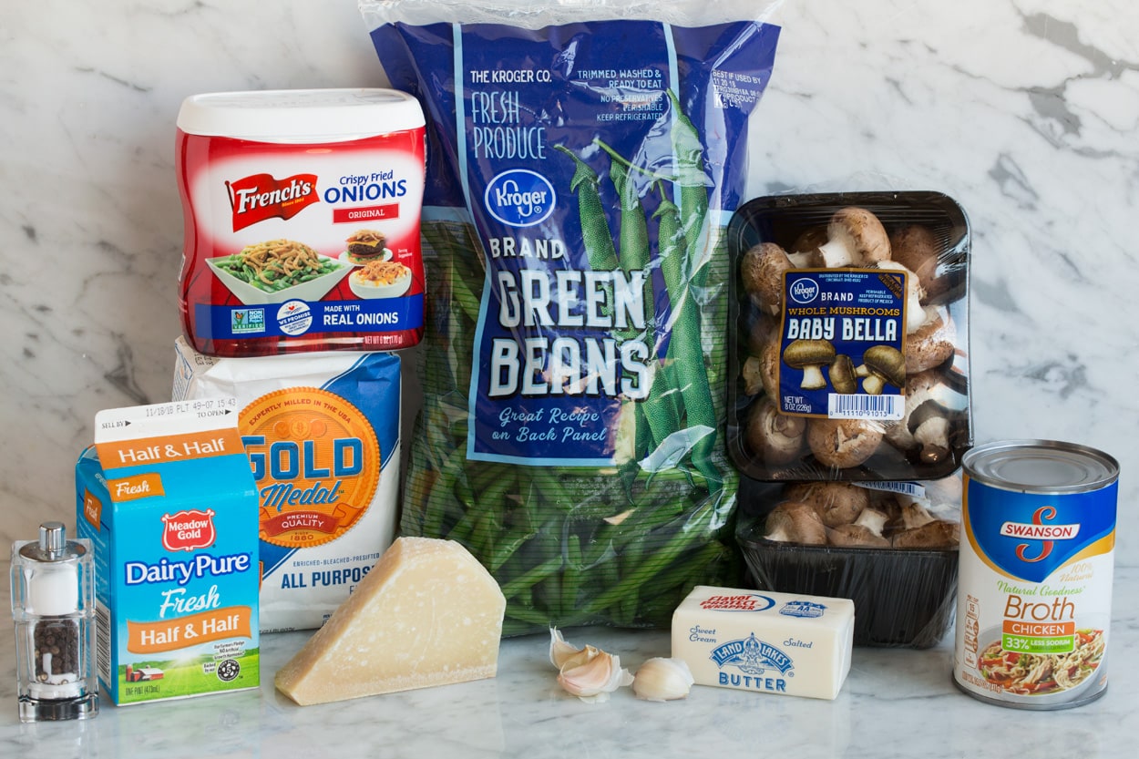 Ingredients for Green Bean Casserole shown here on a white marble surface. They include fresh green beans, mushrooms, French's fried onions, flour, butter, half and half, chicken broth, parmesan, and garlic.
