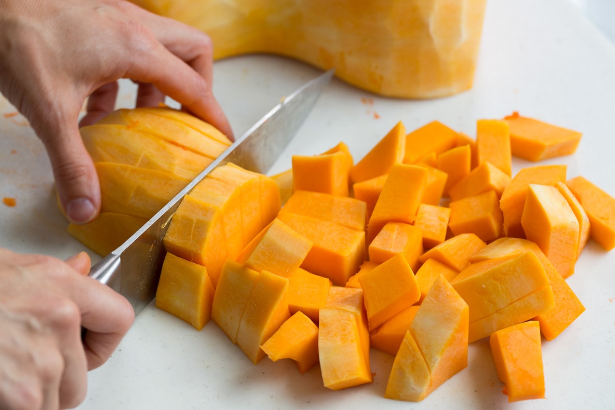 Showing how to cut a whole butternut squash into cubes on a cutting board with a knife.