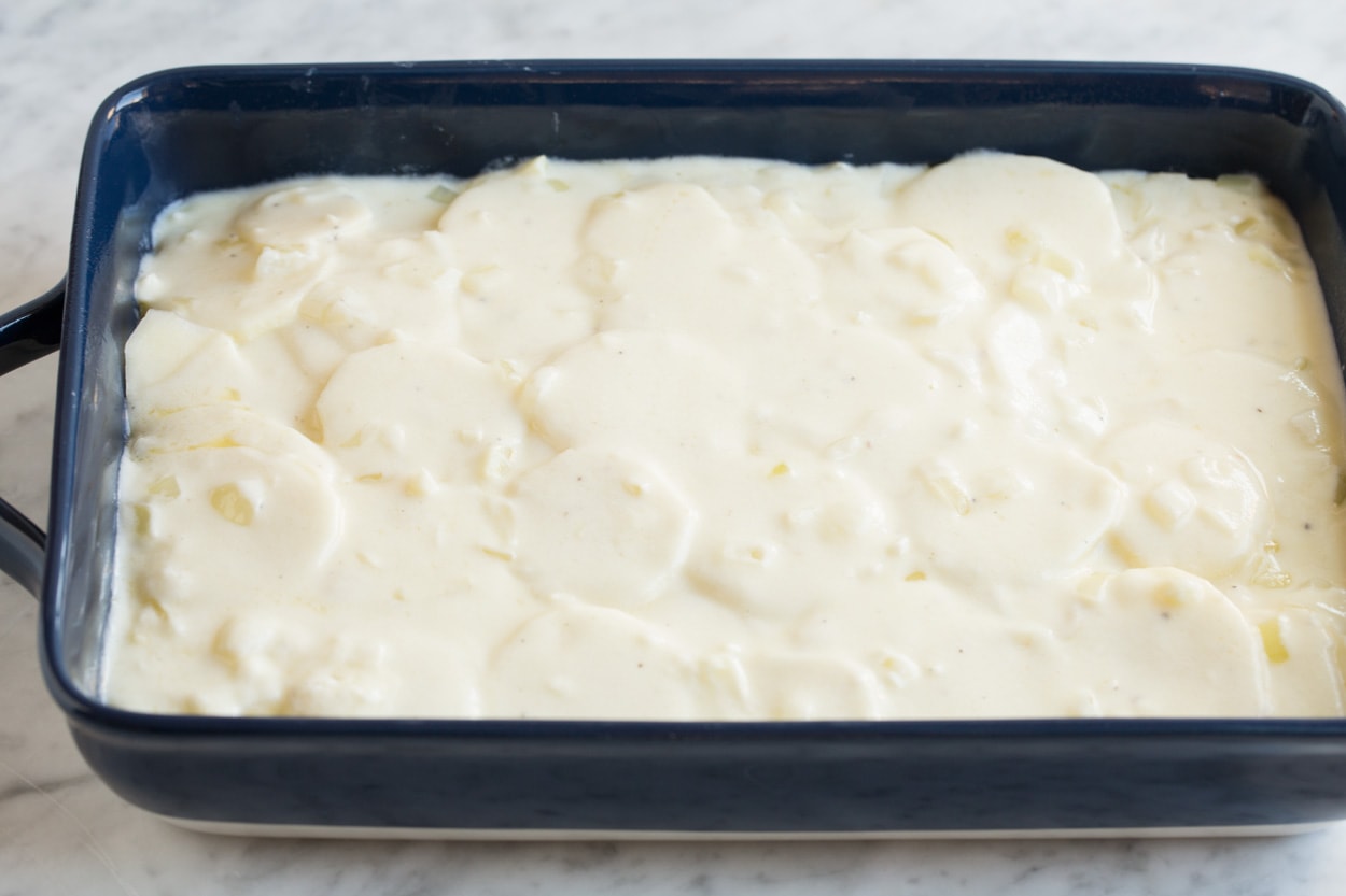 Finishing scalloped potatoes by adding a final layer of sauce over thinly sliced potatoes in baking dish.