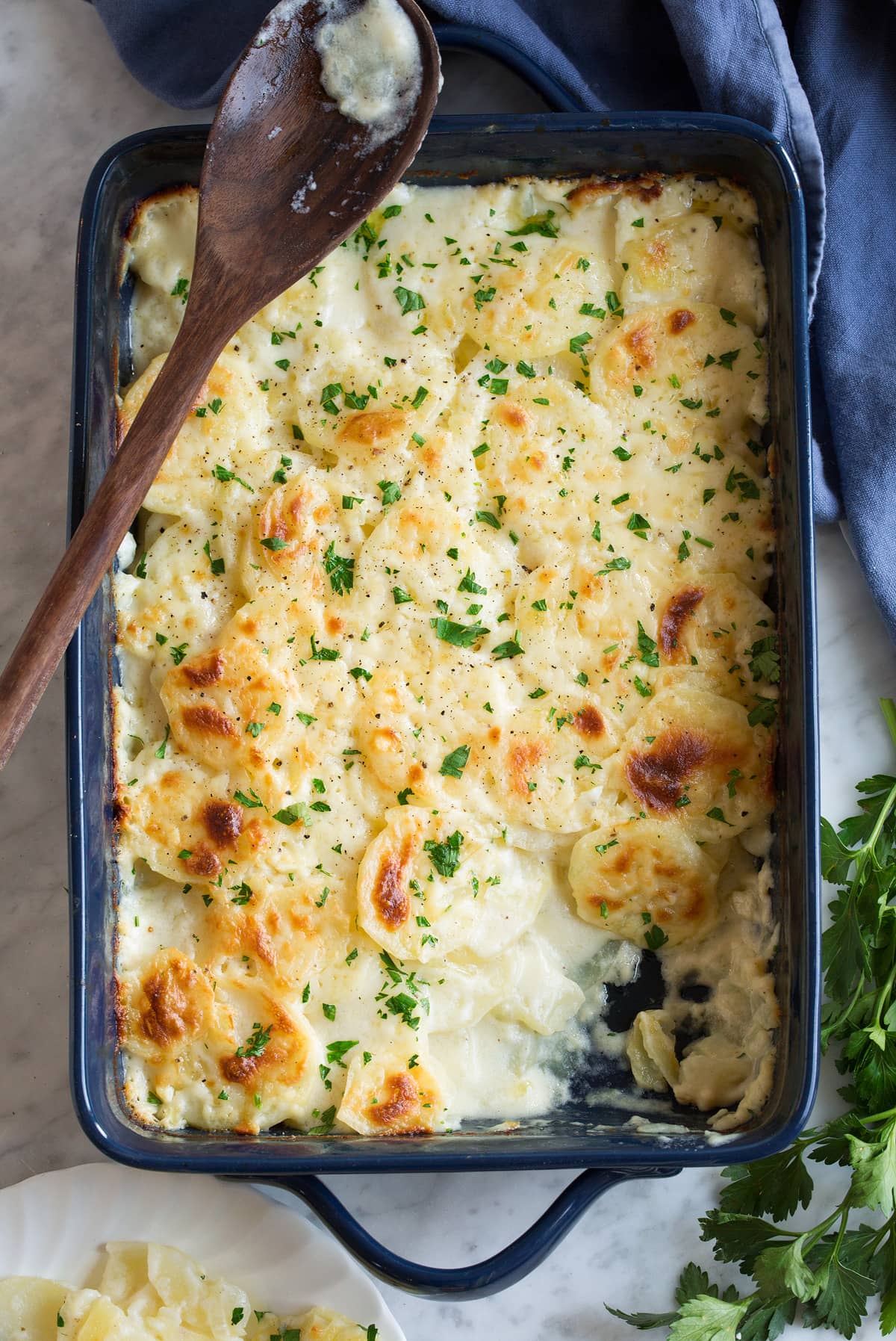Scalloped Potatoes after baking in a dark baking dish set over marble surface. One scoop is removed to show sauce and thin sliced potatoes.
