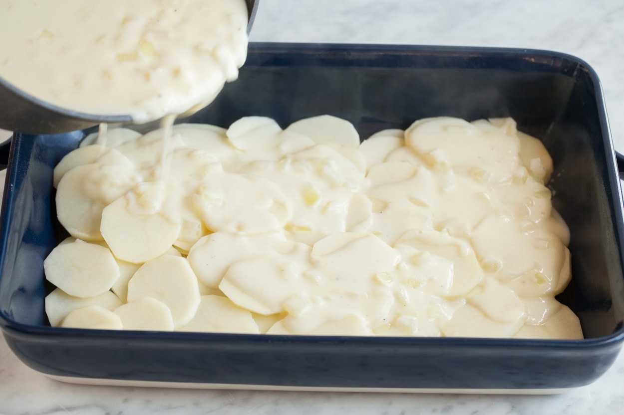 Pouring milk and cheese sauce over thinly sliced potatoes in a baking dish.