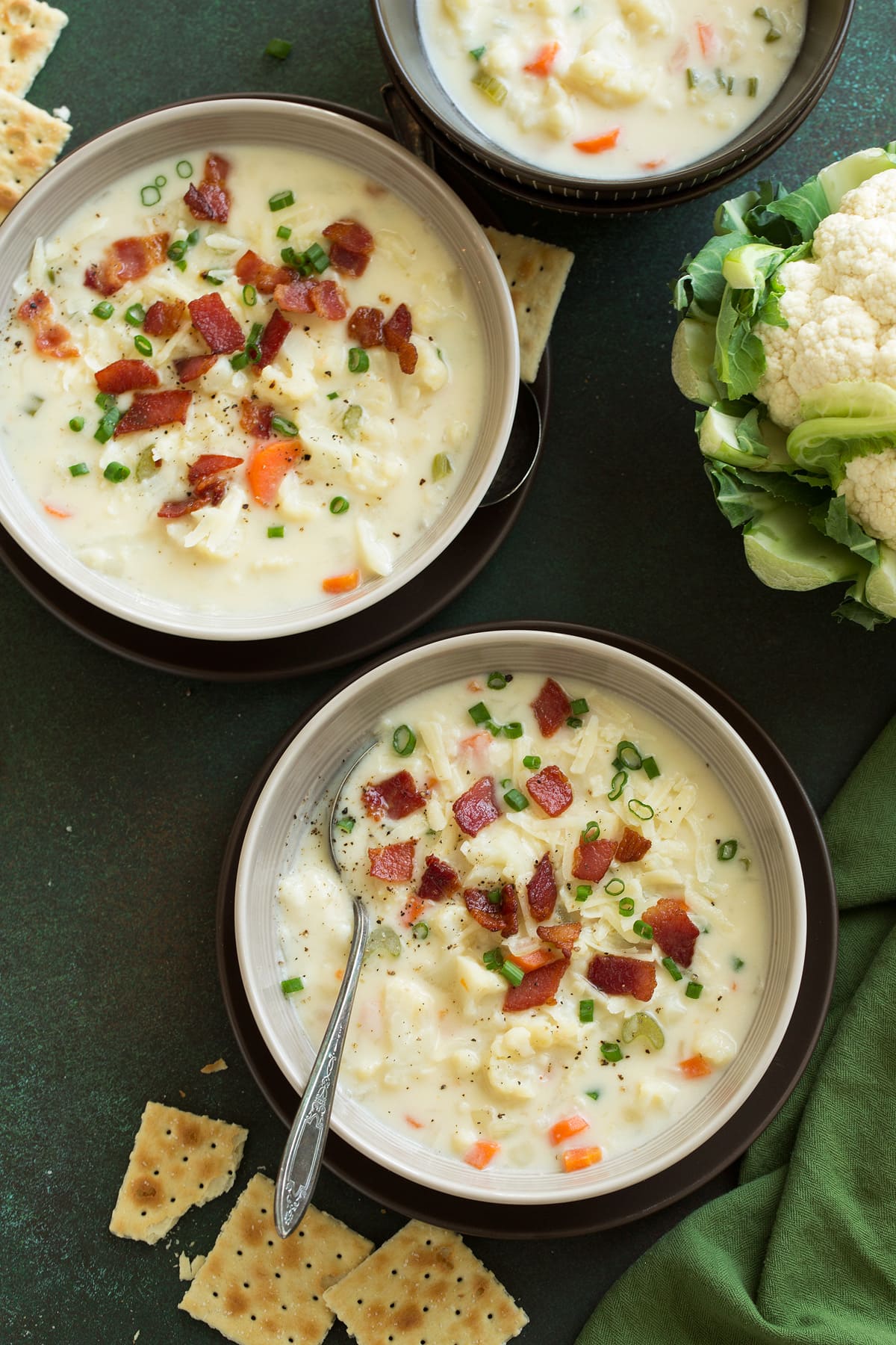 Three servings of Cauliflower Soup in grey bowls set over brown plates on a green tabletop surface.