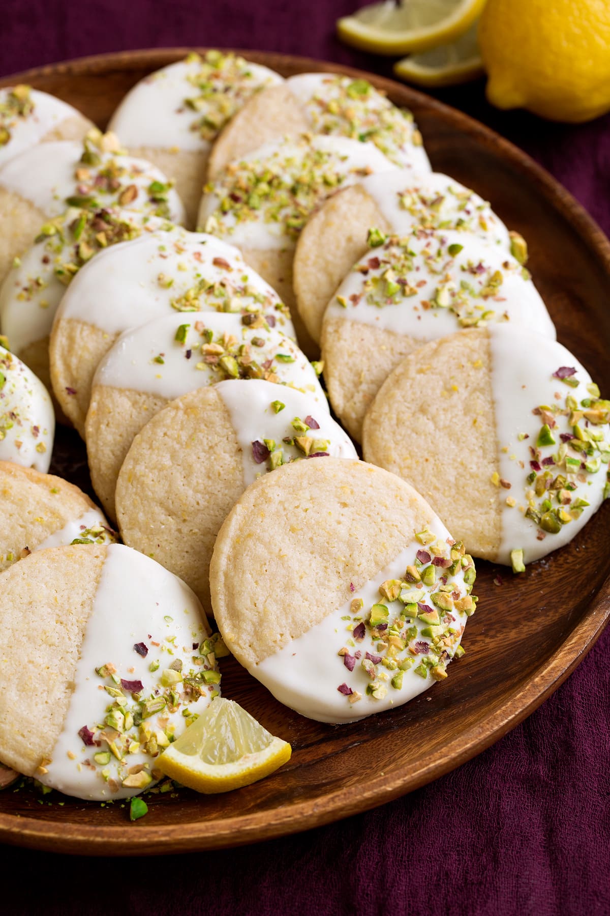 Shortbread cookies with lemon flavor that are dipped in white chocolate and sprinkled with pistachios. Shown here on a wooden serving plate.