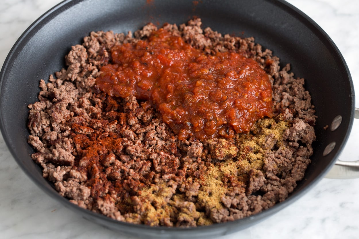 Adding salsa and spices to cooked ground beef mixture in large skillet.