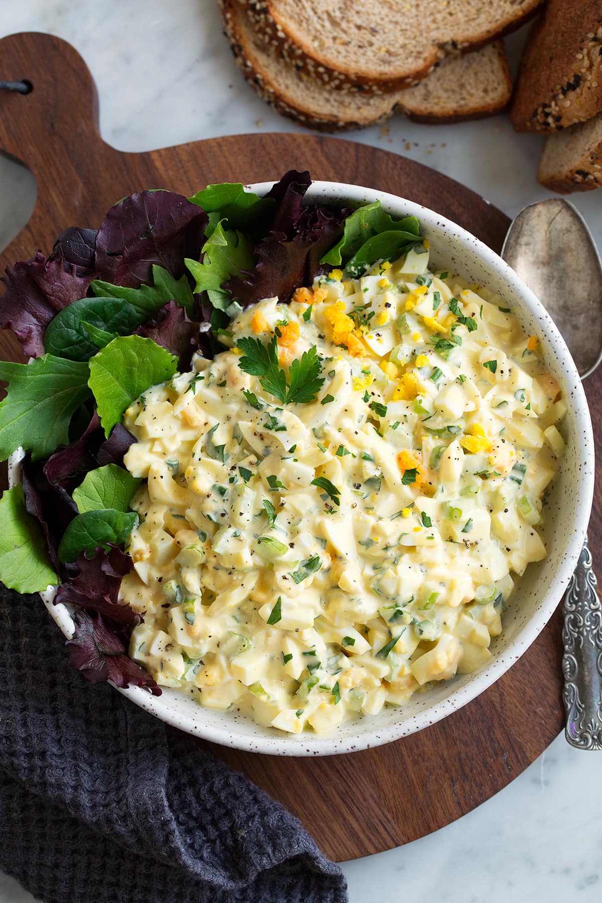 Egg Salad in a white speckled ceramic serving bowl with a side of colorful lettuce. Bowl is sitting on a wooden platter which is on a marble tabletop. There is a silver spoon of the the side and whole grain bread in the corner.