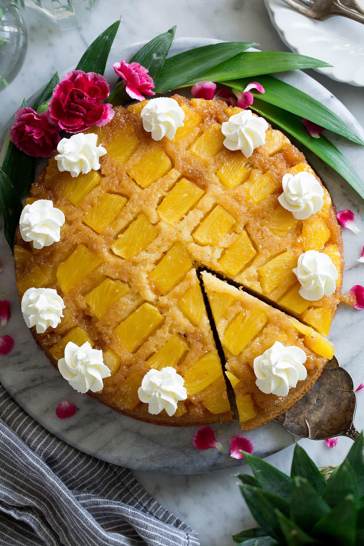 Overhead image of Pineapple Upside Down Cake showing rows of pineapple chunks. Cake is decorated with swirls of whipped cream. Along the edge of the cake are pink flowers and pineapple leaves for decoration.