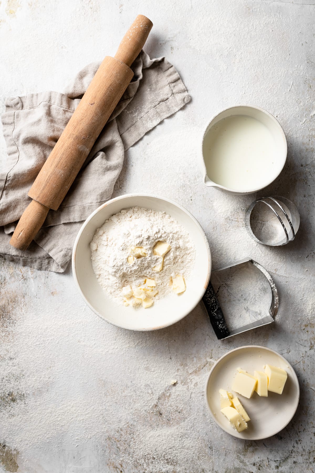 Showing biscuit ingredients including flour mixture, butter and buttermilk in mixing bowls. Rolling pin on the side and beige kitchen towel.