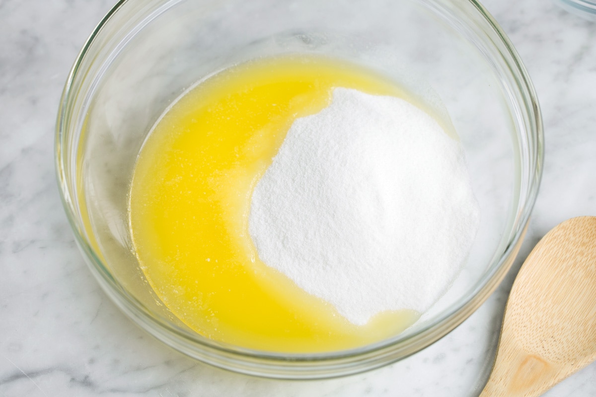 Adding sugar to melted butter in glass mixing bowl for brownies.