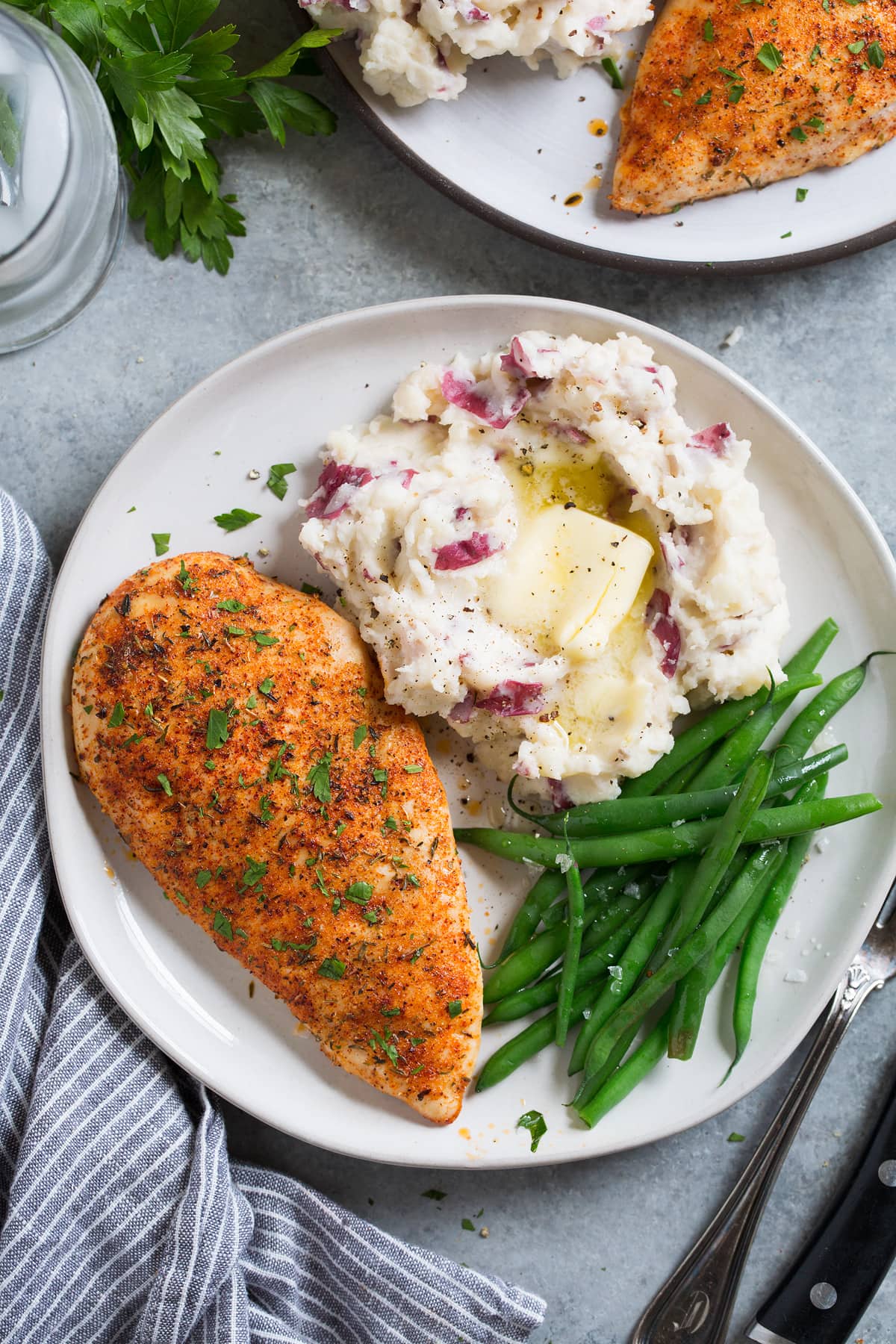 Overhead image of a seasoned baked chicken breast on a white serving plate along with a side of mashed potatoes and steamed green beans.