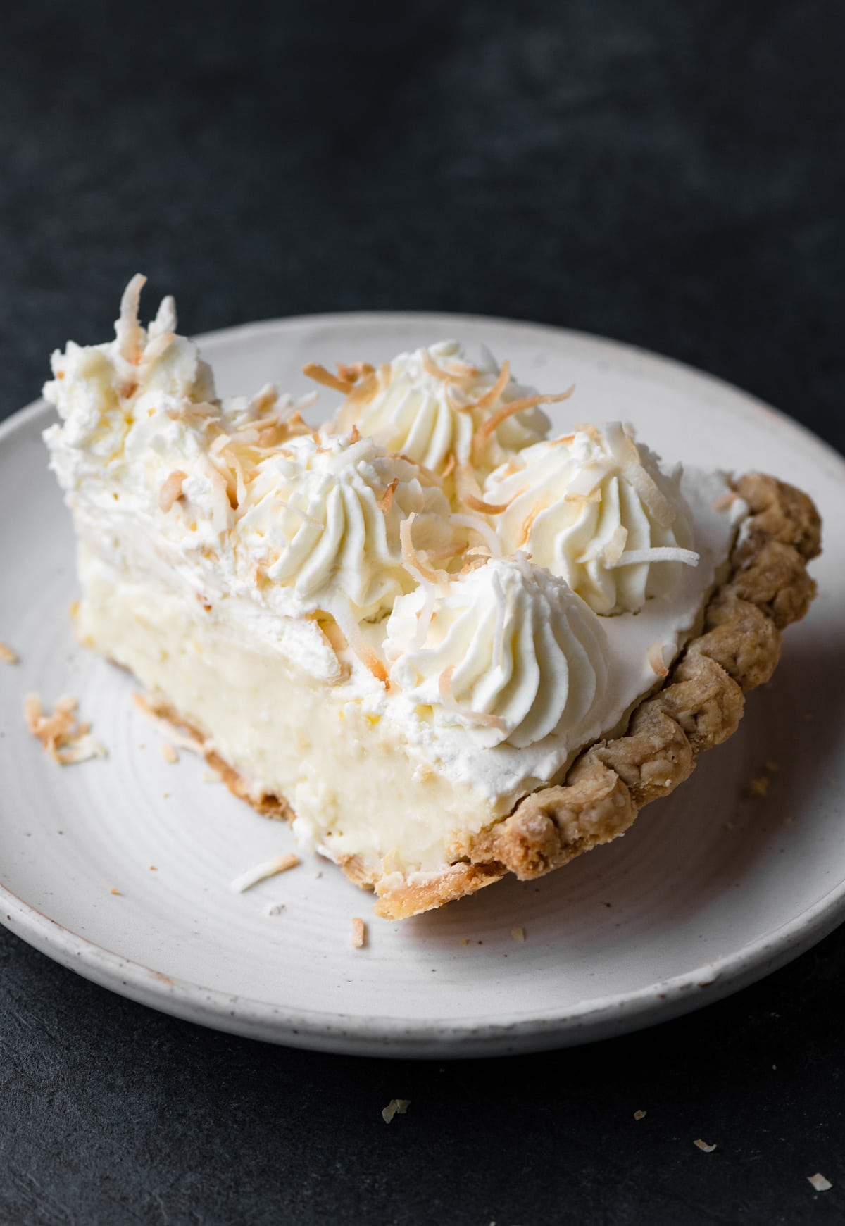 Slice of coconut cream pie on a white plate set over a dark grey tabletop.