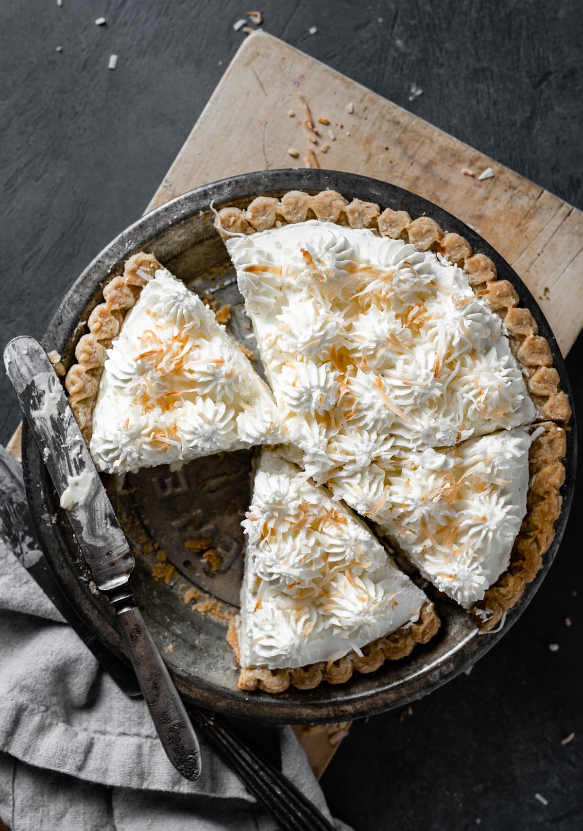 Whole coconut cream pie in a pie dish with slices cut. Pie is sitting on a wooden cutting board over a dark surface.