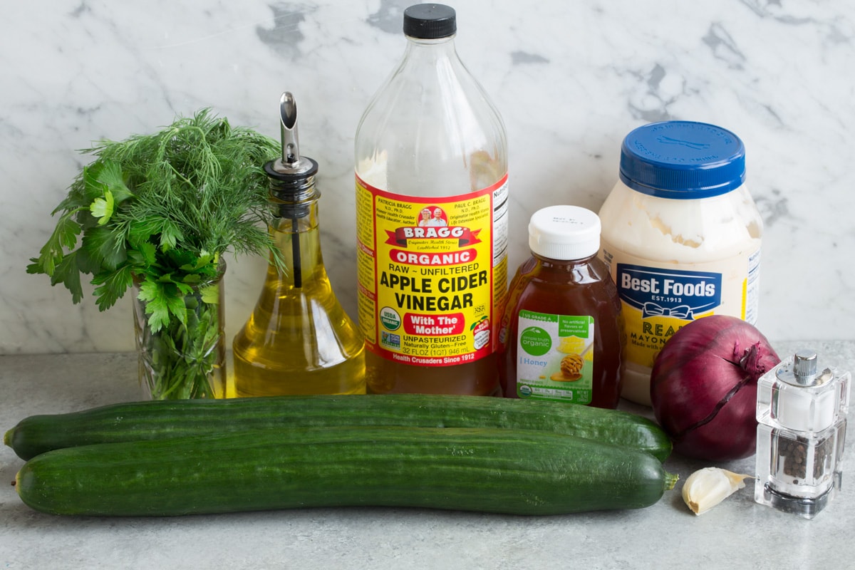 Ingredients needed to make cucumber salad shown here including English cucumbers, mayonnaise, olive oil, garlic, apple cider vinegar, dill, parsley, honey, and red onion.