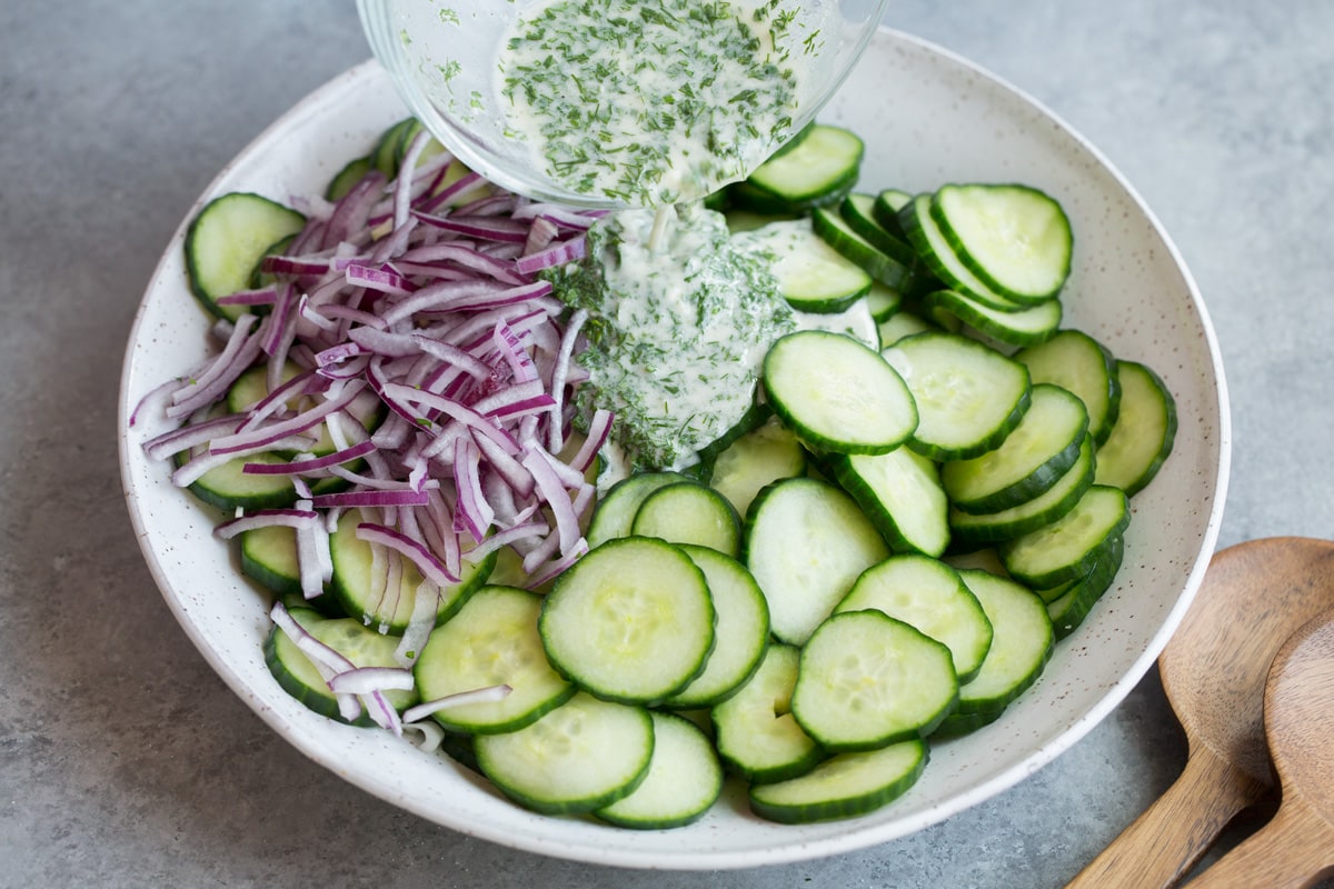 Pouring dressing over cucumbers and red onion in white serving bowl.