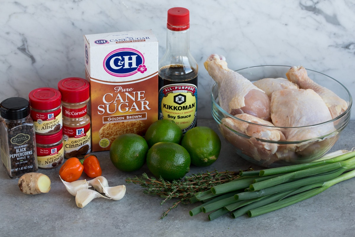 Jerk Chicken ingredients shown here including chicken pieces, green onions, thyme, limes, soy sauce, brown sugar, cinnamon, allspice, nutmeg, pepper, scotch bonnet pepper, garlic, and fresh ginger.