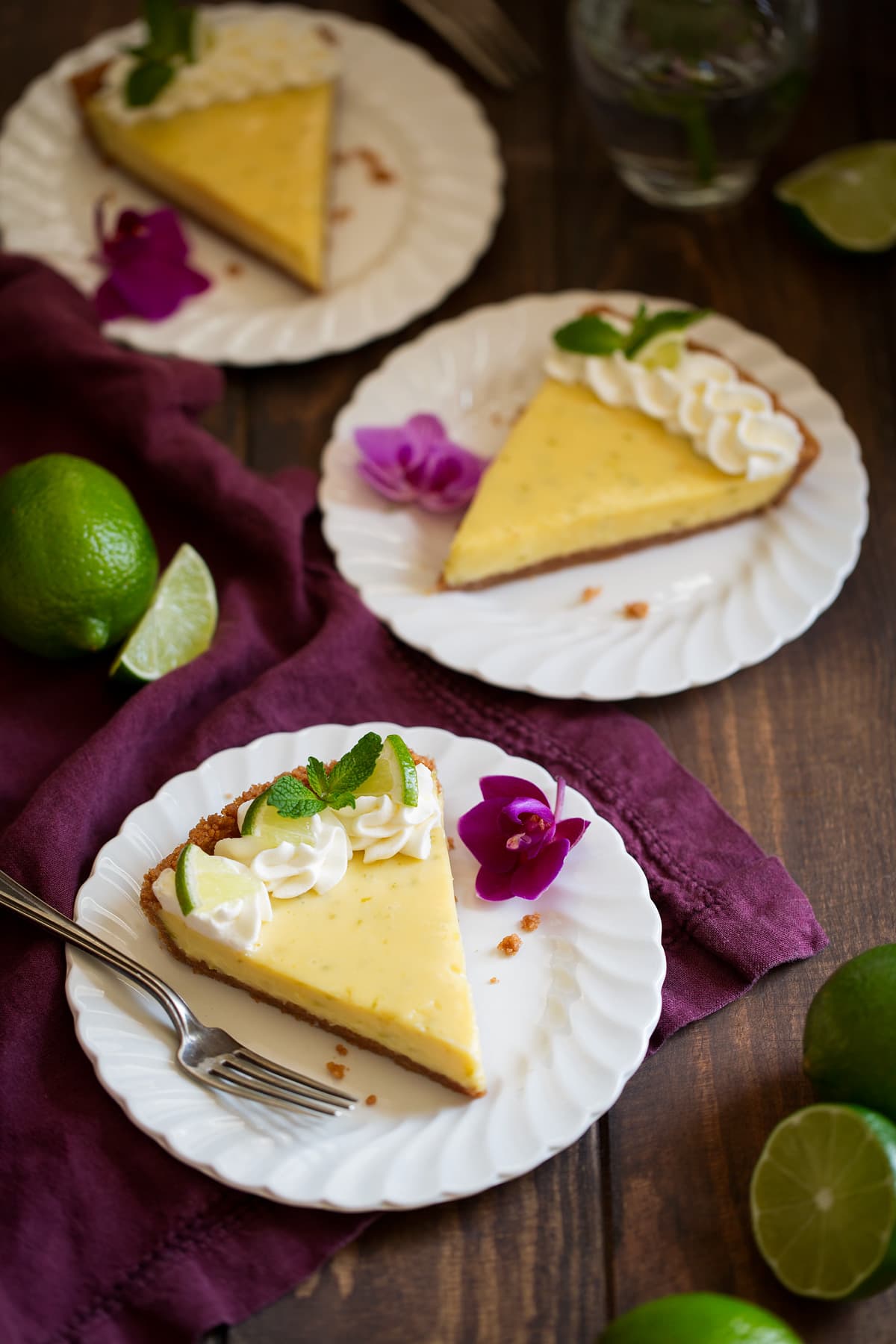 Three slices of key lime pie on white scalloped dessert plates set over a maroon linen napkin. Plates are decorated with purple orchids.
