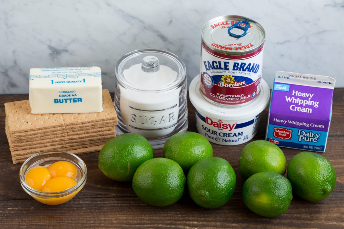 Eight ingredients needed for key lime pie shown here including graham crackers, butter, sugar, egg yolks, limes, sour cream, sweetened condensed milk and heavy cream.