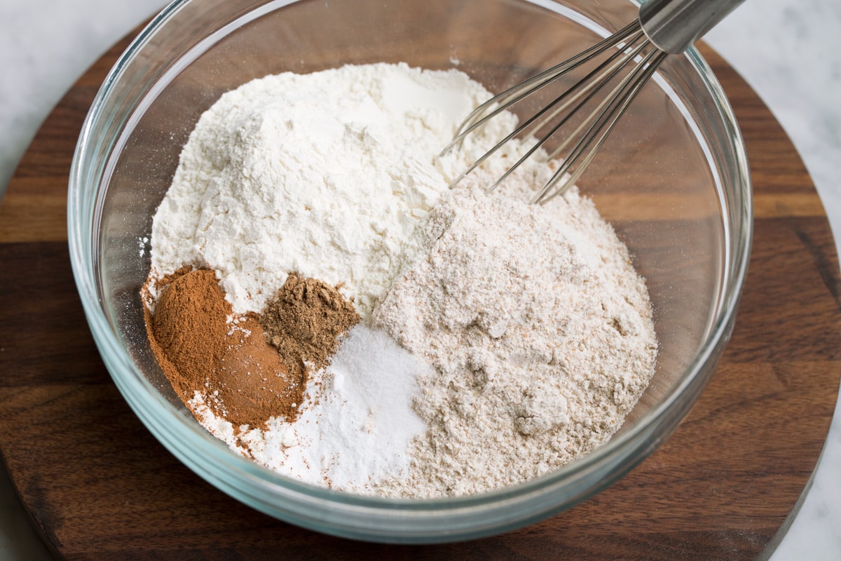 Showing how to make morning glory muffins. Whisking white flour, wheat flour, cinnamon, nutmeg, baking soda and salt in a glass mixing bowl.