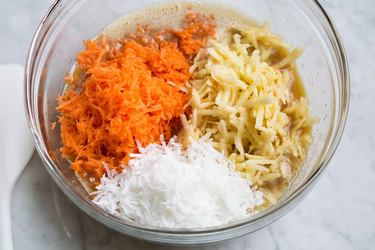 Adding shredded carrots, apples and coconut to morning glory muffin batter in glass mixing bowl.