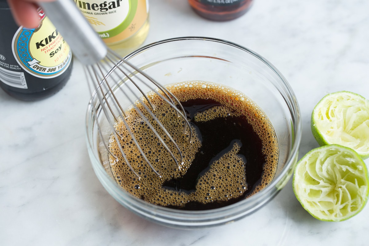 Showing how to make pad thai sauce. Whisking soy sauce, fish sauce, lime juice, rice vinegar and brown sugar in a glass mixing bowl set over a marble surface.