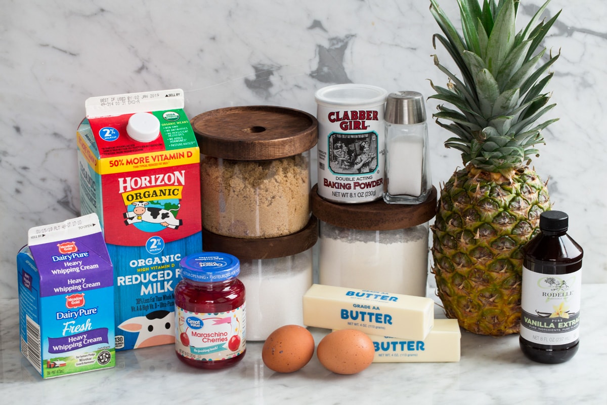 Ingredients needed to make pineapple upside down cake shown here including fresh pineapple, flour, brown and white sugar, butter, eggs, baking powder, salt, milk, cream and maraschino cherries.