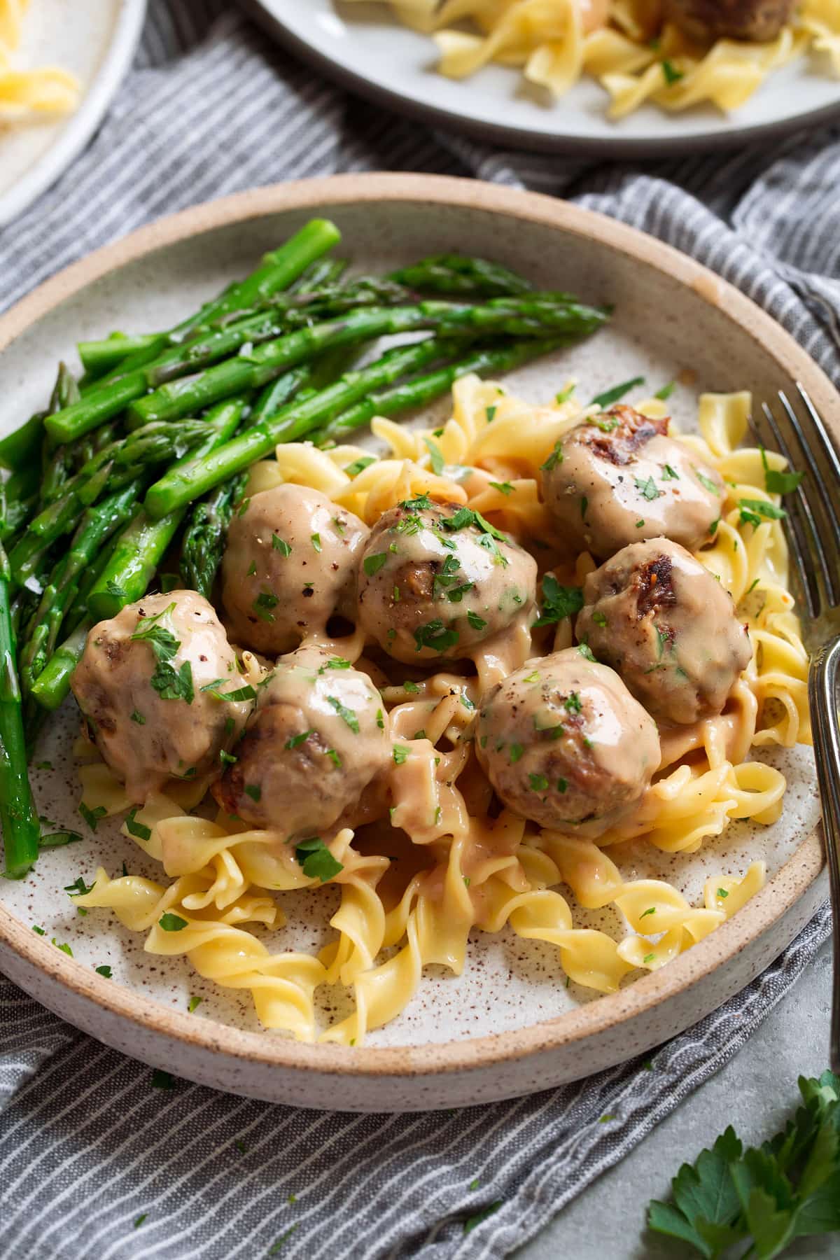 Serving of Swedish meatballs on egg noodles with a side of asparagus on a speckled cream plate.