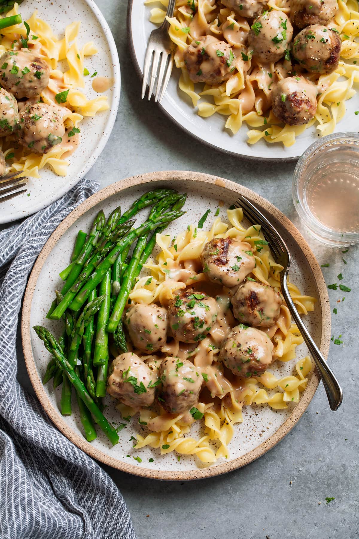 Three servings of Swedish meatballs on plates. Meatballs are laying over egg noodles and served with a side of steamed asparagus.