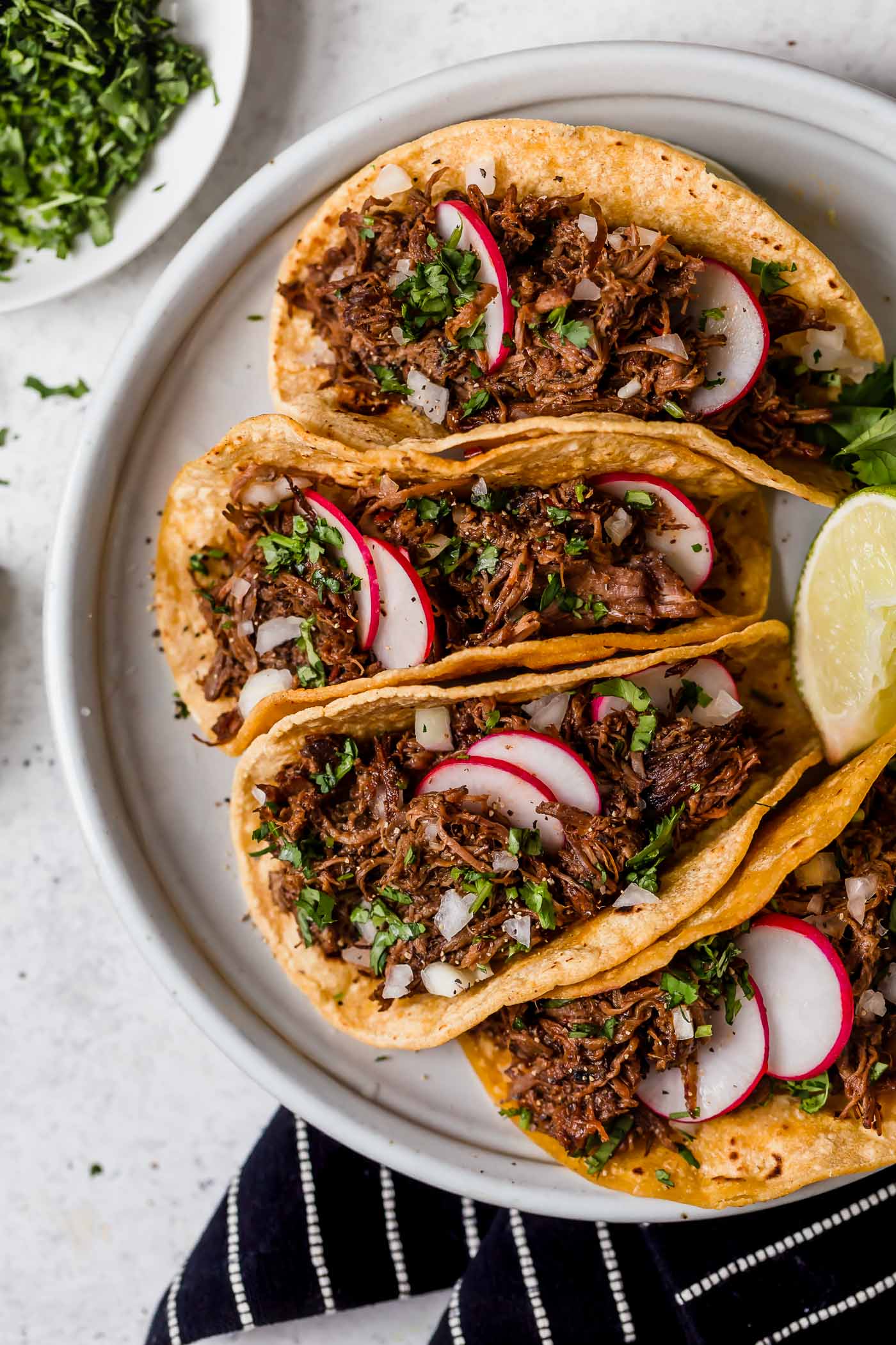Barbacoa Beef Barbacoa beef layered in corn tortillas to make tacos. Shown sitting on a light grey serving platter, beef is garnished with cilantro and radishes.
