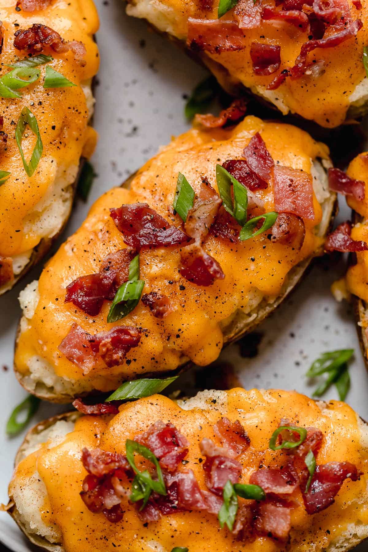 Overhead image of four Twice Baked Potatoes on a white serving plate. Potato skins are filled with mashed potatoes, topped with melted cheddar cheese and garnished with bacon and green onions. Close up image of twice baked potatoes on a serving plate.