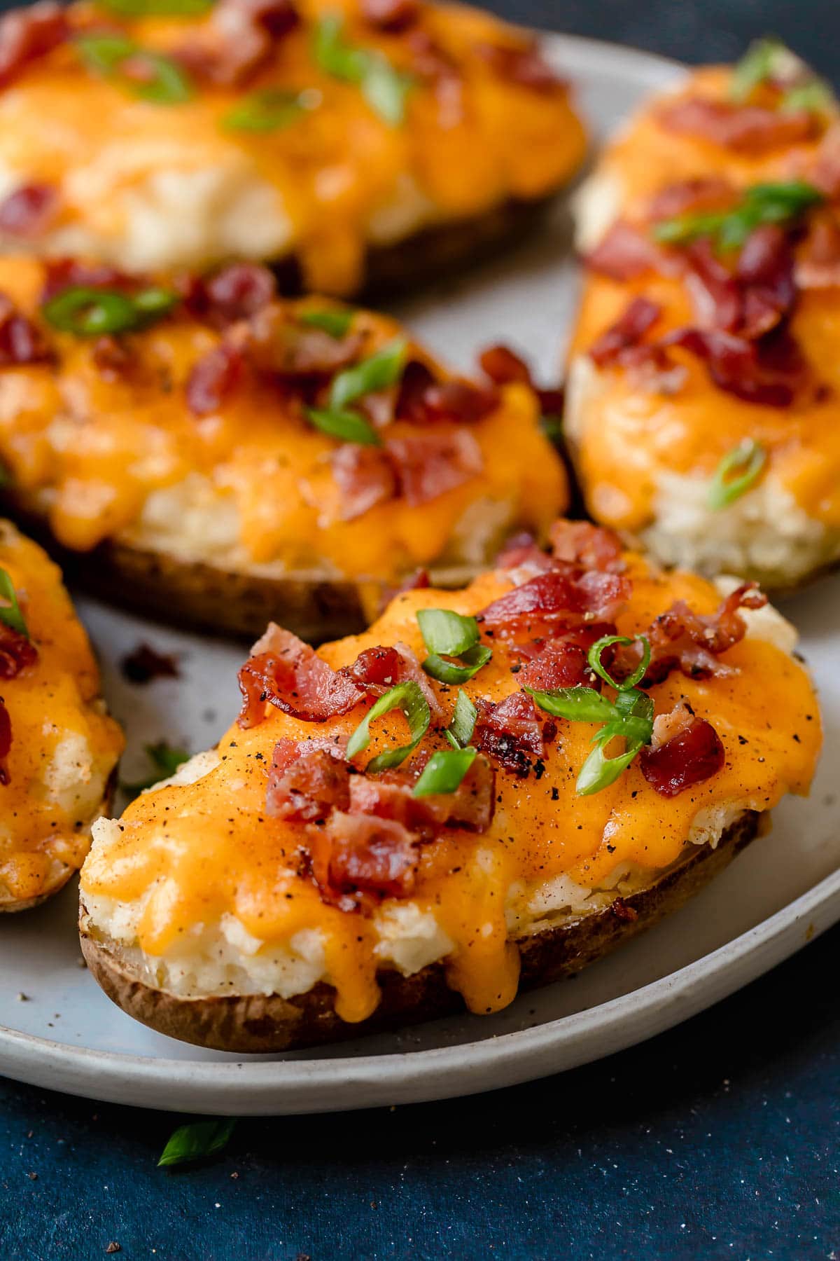 Overhead image of four Twice Baked Potatoes on a white serving plate. Potato skins are filled with mashed potatoes, topped with melted cheddar cheese and garnished with bacon and green onions. Side view of a filled twice baked potato.