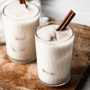 Horchata in two glass cups with ice cubes and a cinnamon stick. Cups are set over a wooden cutting board.