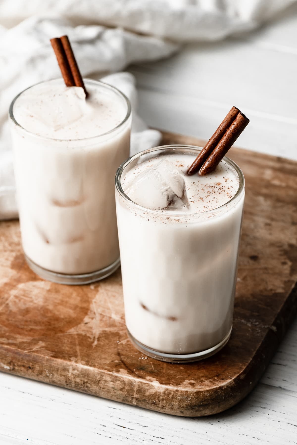 Horchata in two glass cups with ice cubes and a cinnamon stick. Cups are set over a wooden cutting board.