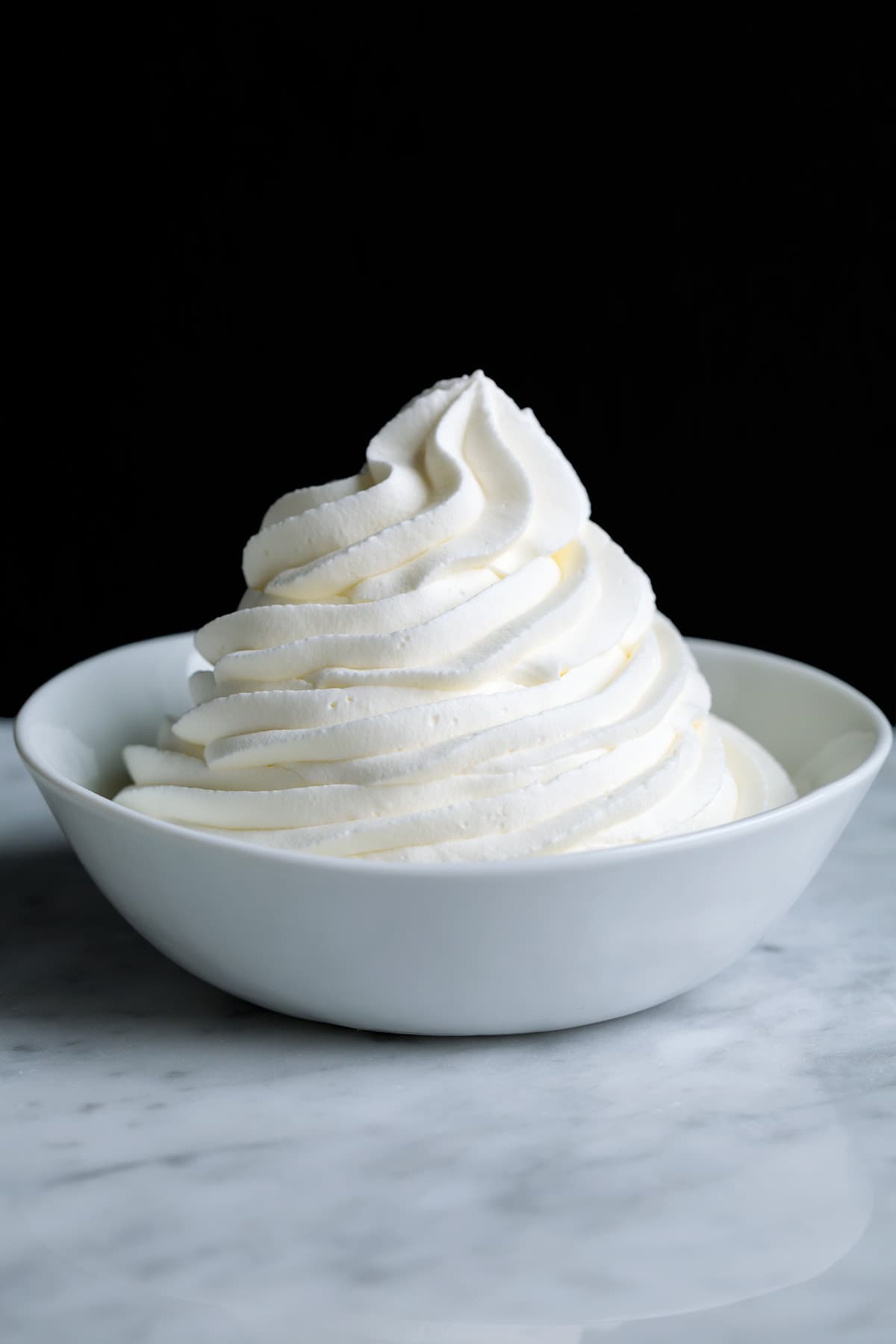 Piped mound of whipped cream in a white bowl set over a marble surface with a black background.