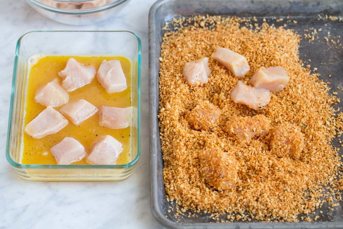 Showing how to make chicken nuggets. Tossing chicken pieces with egg then dressing in panko breadcrumb mixture on baking sheet.