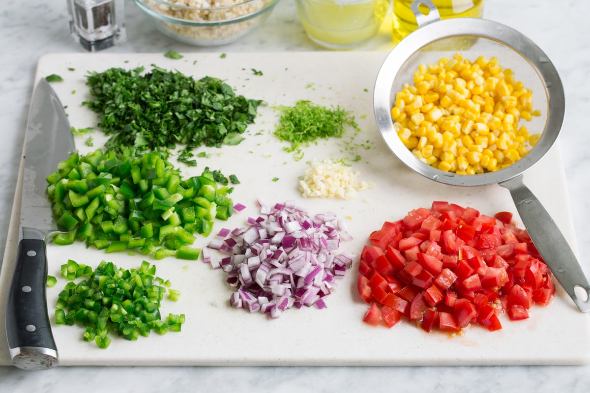 Chopped vegetables on a cutting board with corn in a colander to drain and rice in the background in a bowl.